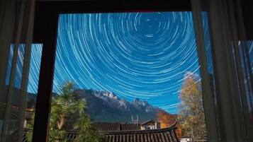 Vista a través de una ventana que muestra un paisaje montañoso, con arquitectura tradicional y un cielo nocturno despejado con estelas de estrellas.