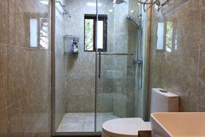 Una ducha con mampara de vidrio, un cabezal de ducha manual, una ventana pequeña y un inodoro blanco.