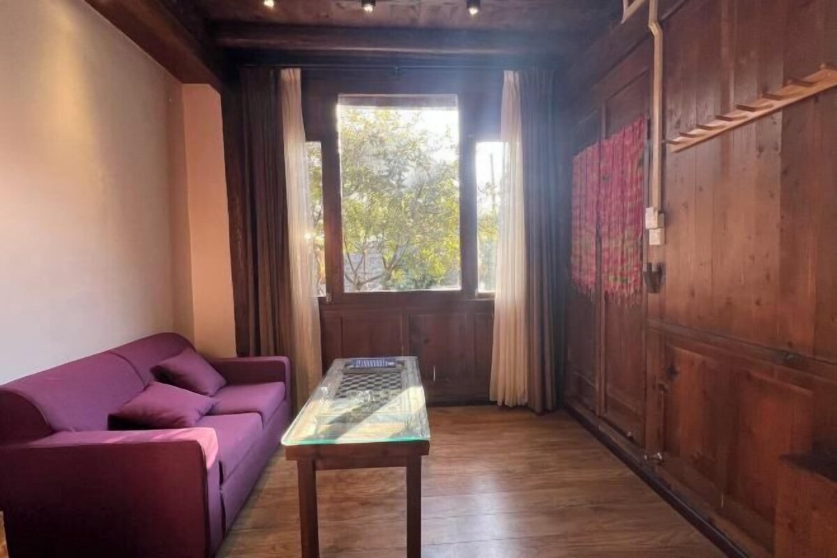 Una habitación con un sofá morado, una mesa de vidrio, piso de madera, un ventanal con cortinas y una puerta de madera.