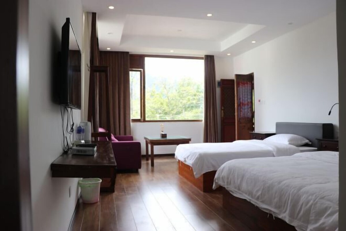 Habitación de hotel con dos camas, un escritorio con silla, televisor montado en la pared y ventana con vista a la vegetación.