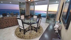 A modern living room with a glass table and chairs, a patterned sofa, and a view of the ocean at sunset.