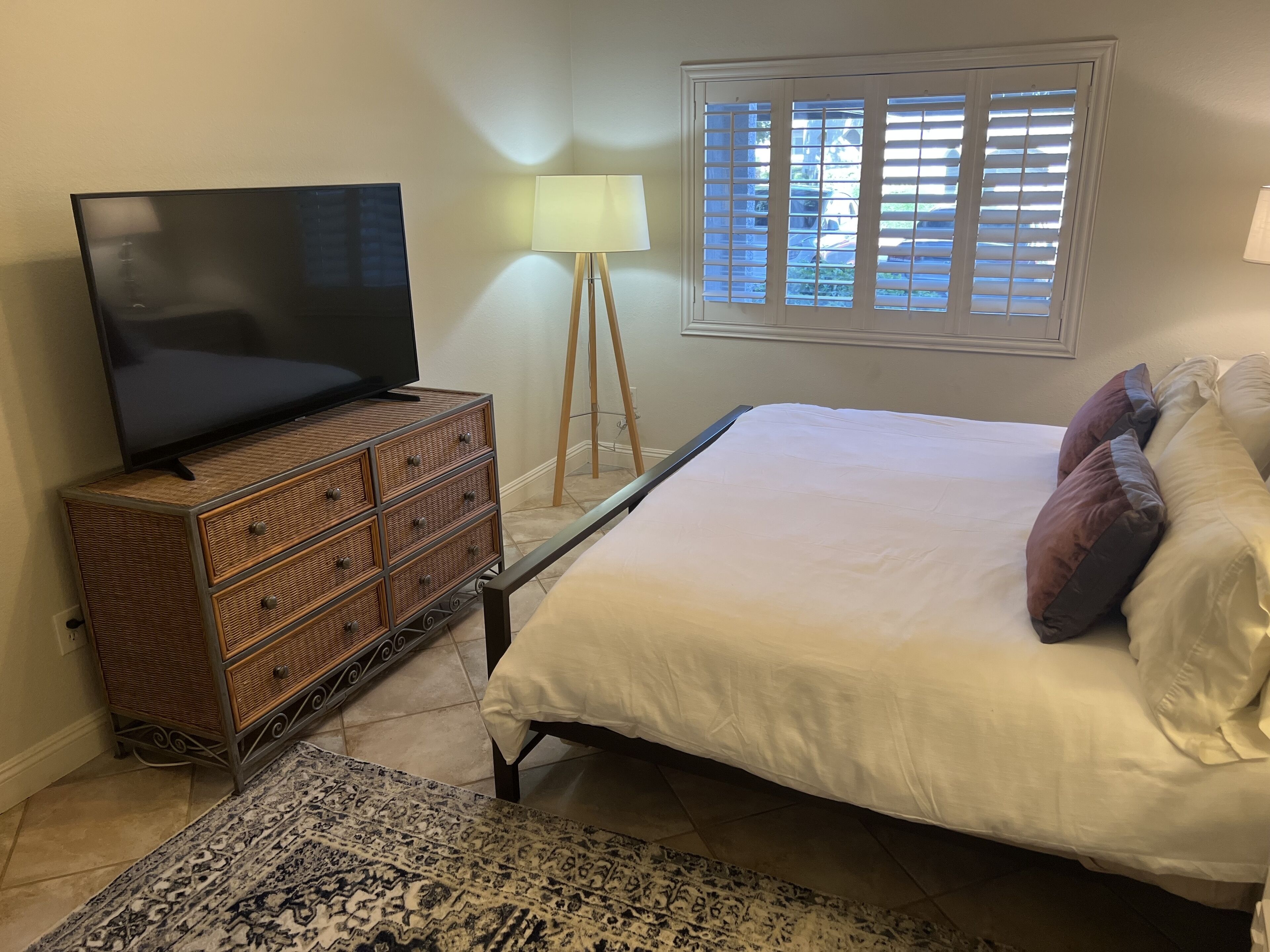 A bedroom with a bed, a TV on a dresser, a lamp, and a window with blinds.