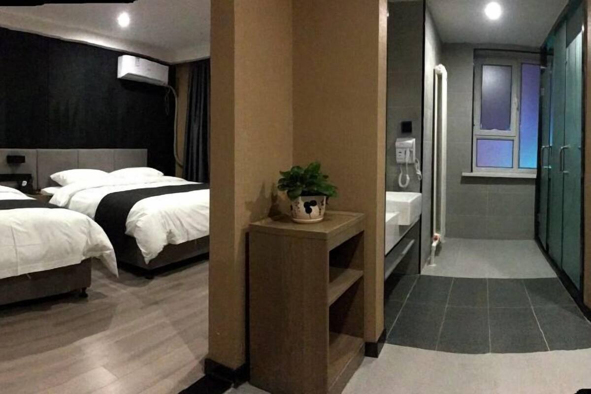 Ein Hotelzimmer mit zwei Betten, einem kleinen Holfnachtkästchen mit Pflanze, einem Badezimmer mit Dusche und einem Flur, der zu einem weiteren Zimmer führt.