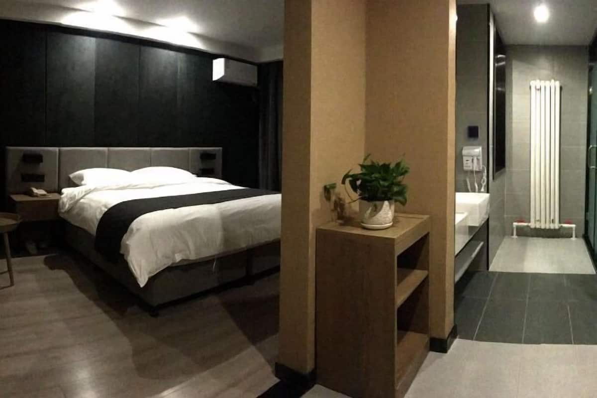 Ein modernes Hotelzimmer mit einem großen Bett, einem Nachttisch mit Pflanze, einem kleinen Tisch und einem Badezimmer mit Dusche und Toilette.