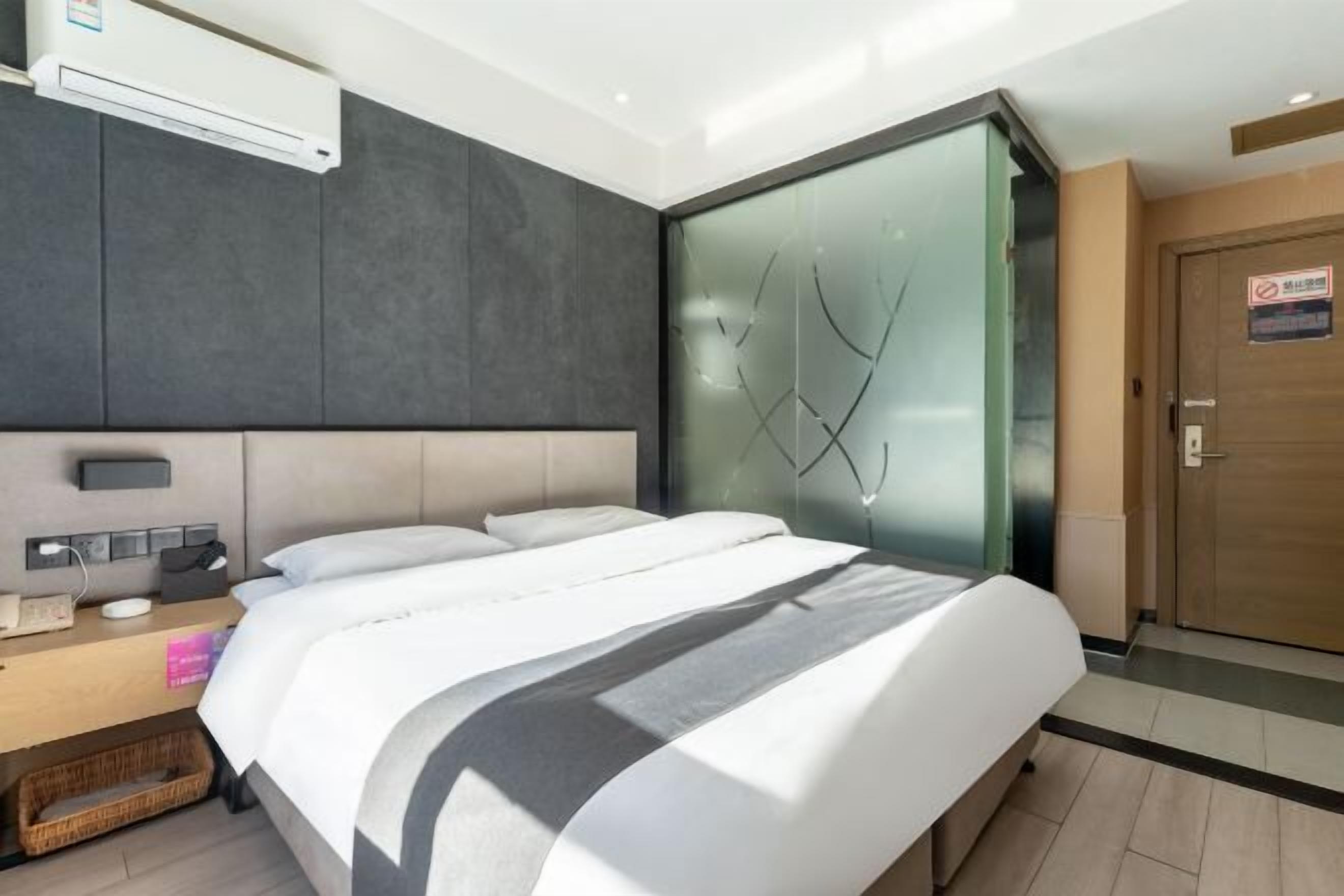 A modern hotel room with a large bed, a bedside table with a lamp, and a wall-mounted air conditioner.
