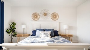 Ein Schlafzimmer mit einem weißen Bett, blau-weißer Bettwäsche, zwei Nachttischen mit Lampen, einer Wand mit drei dekorativen Wandbildern und einer Topfpflanze.