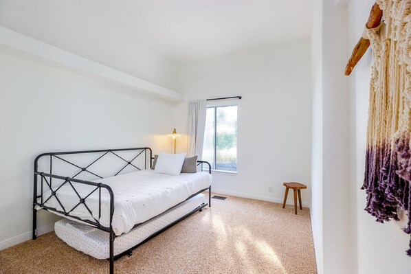 A bedroom with a metal bed frame, a white mattress, a bedside lamp, a window with curtains, and a small stool. - 25% Off Oct 29-31, Perfect For Families, W/Pool! (Temecula)