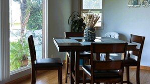 A dining area with a wooden table, four chairs, a woven pendant light, a vase with dried plants, and a window with a view of greenery. - Cozy Escape - Beach Community of Playa Del Rey (Los Angeles)