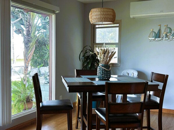 A dining area with a wooden table, four chairs, a woven pendant light, a vase with dried plants, and a window with a view of greenery. - Cozy Escape - Beach Community of Playa Del Rey (Los Angeles)