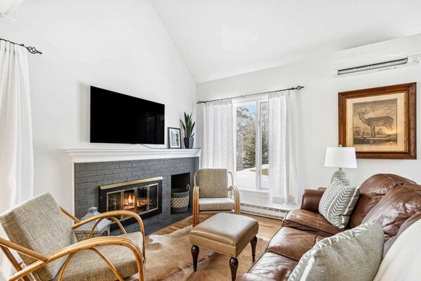 A living room with a fireplace, a television mounted above it, wicker chairs, a leather sofa, and a large window with curtains.