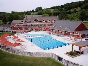 A large swimming pool with multiple lanes, surrounded by lounge chairs and umbrellas, with a building featuring a red facade and a balcony in the background. - Highland Golf Course Views! - Fairway Village 3A (Quechee)