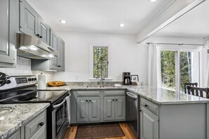 A compact kitchen with grey cabinets, a built-in oven, a stove, and a dishwasher. There is a window providing a view of trees outside. - Highland Golf Course Views! - Fairway Village 3A (Quechee)