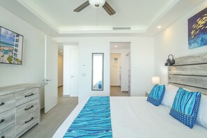 A bedroom with a bed, a dresser, a ceiling fan, a painting, and a view of a building with a pool.
