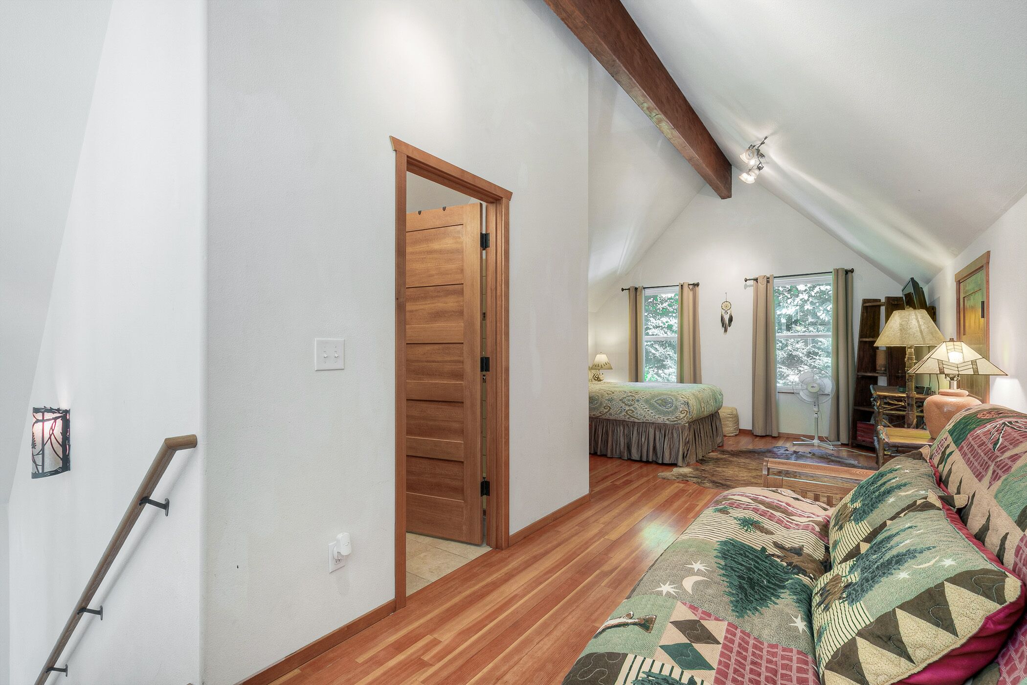 A spacious loft bedroom with a wooden ceiling, a large bed, a sitting area with a patterned sofa, and a view of the outdoors through a window.