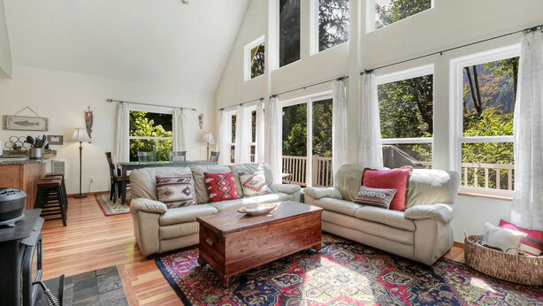 A spacious living room with a beige sofa, a wooden coffee table, and a colorful rug. There are large windows with curtains, a dining area with a table and chairs, and a kitchen area with a stove and utensils.