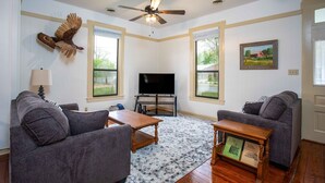 A living room with two grey sofas, a wooden coffee table, a flat-screen TV, and a ceiling fan. - Windmill House | Hot Tub | Great Backyard (Fredericksburg)