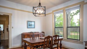 A dining room with a wooden table and chairs, a chandelier, and large windows with a view of trees. - Windmill House | Hot Tub | Great Backyard (Fredericksburg)