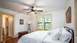 A bedroom with a bed, a ceiling fan, a floor lamp, and a window with a view of trees. - Windmill House | Hot Tub | Great Backyard (Fredericksburg)