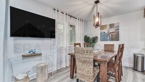 Un comedor con mesa de madera, sillas de mimbre, televisor montado en la pared y una planta sobre la mesa.