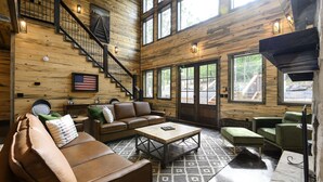 Ein geräumiges Blockhaus-Interieur mit Treppe, Ledersofas, einem Couchtisch und großen Fenstern mit Blick auf den umliegenden Wald.