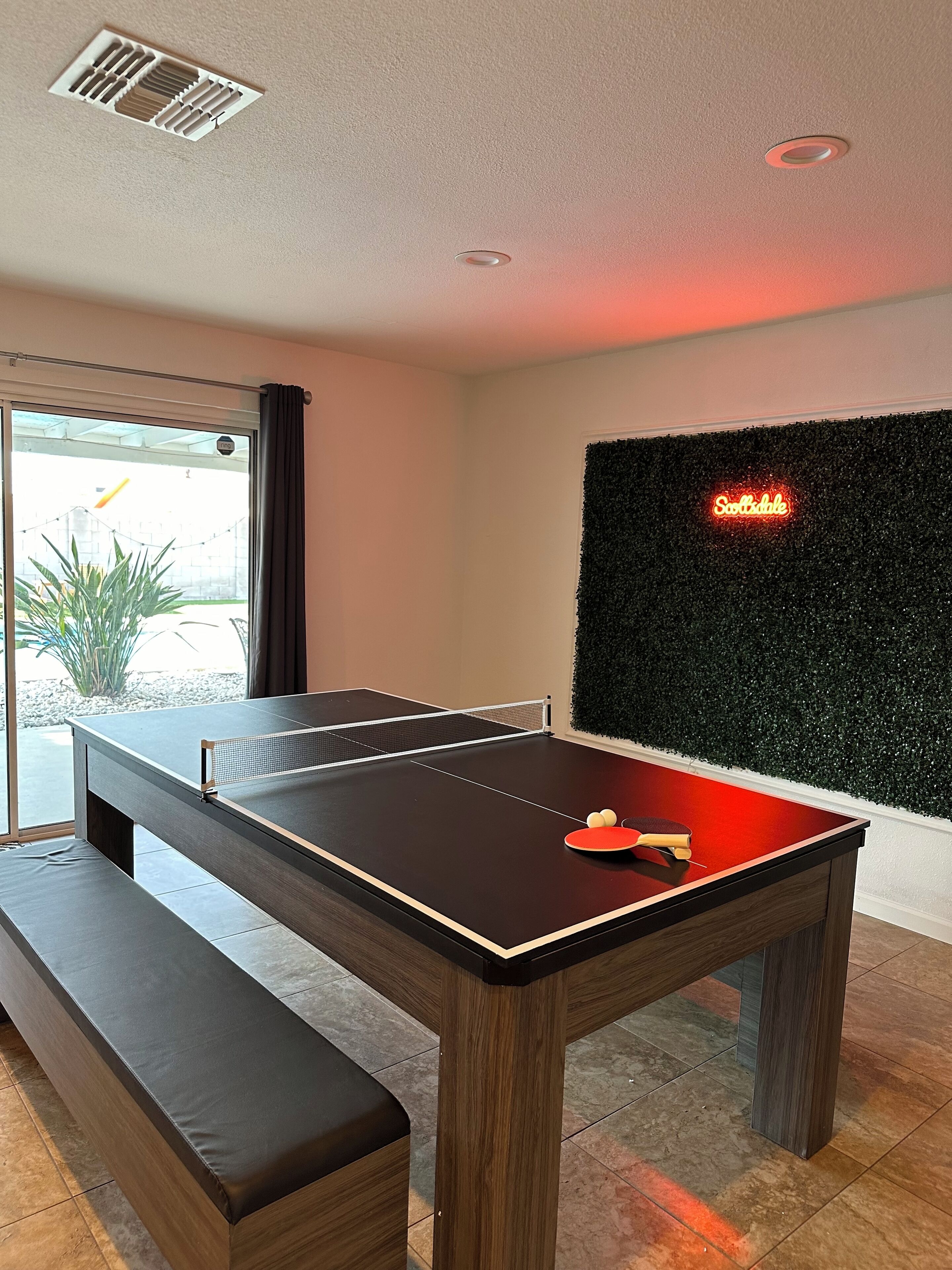 A ping pong table with a bench, a neon sign, and a green wall.
