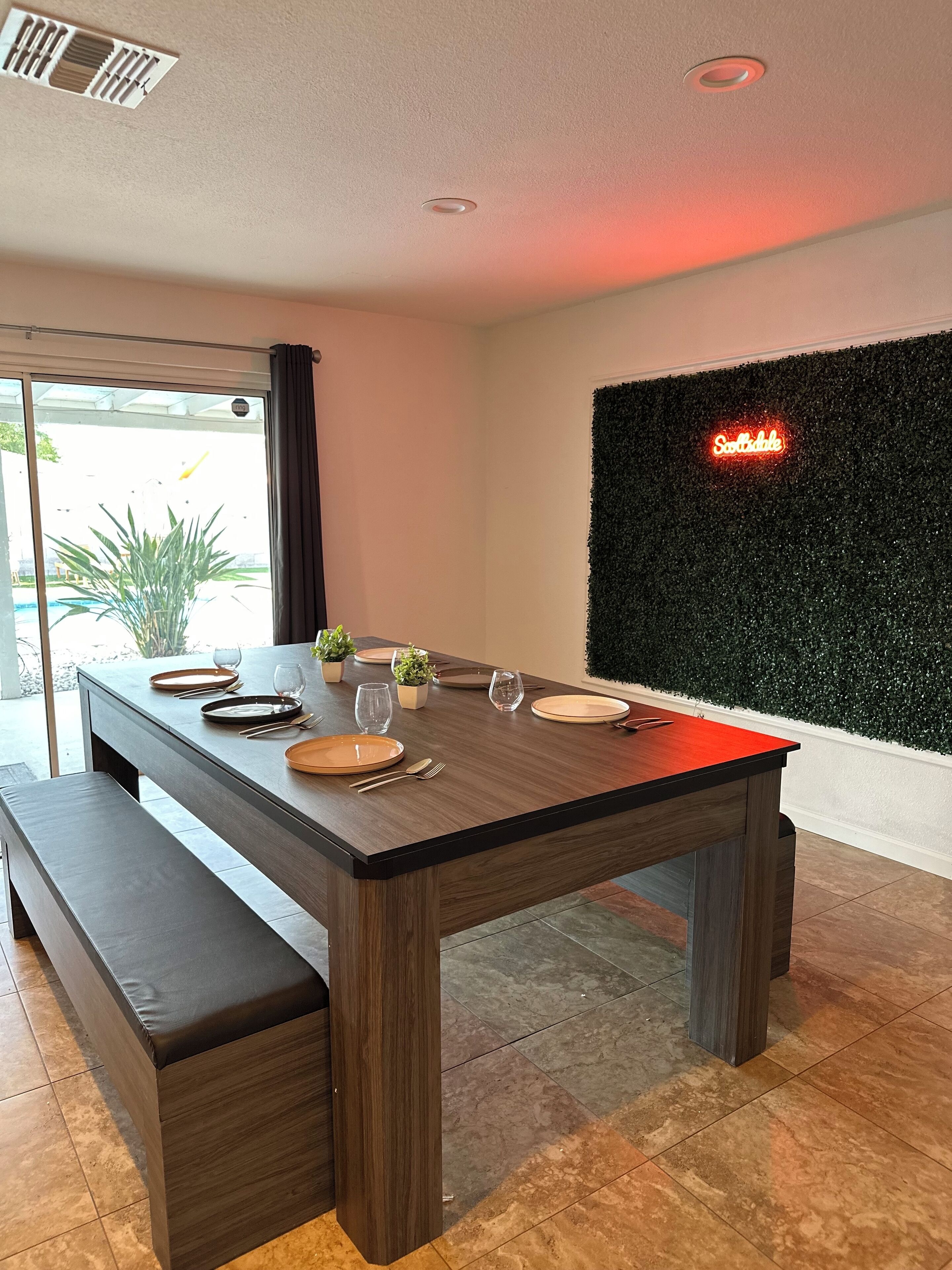 A modern dining area with a wooden table set for four, a bench, and a green wall with a neon sign.