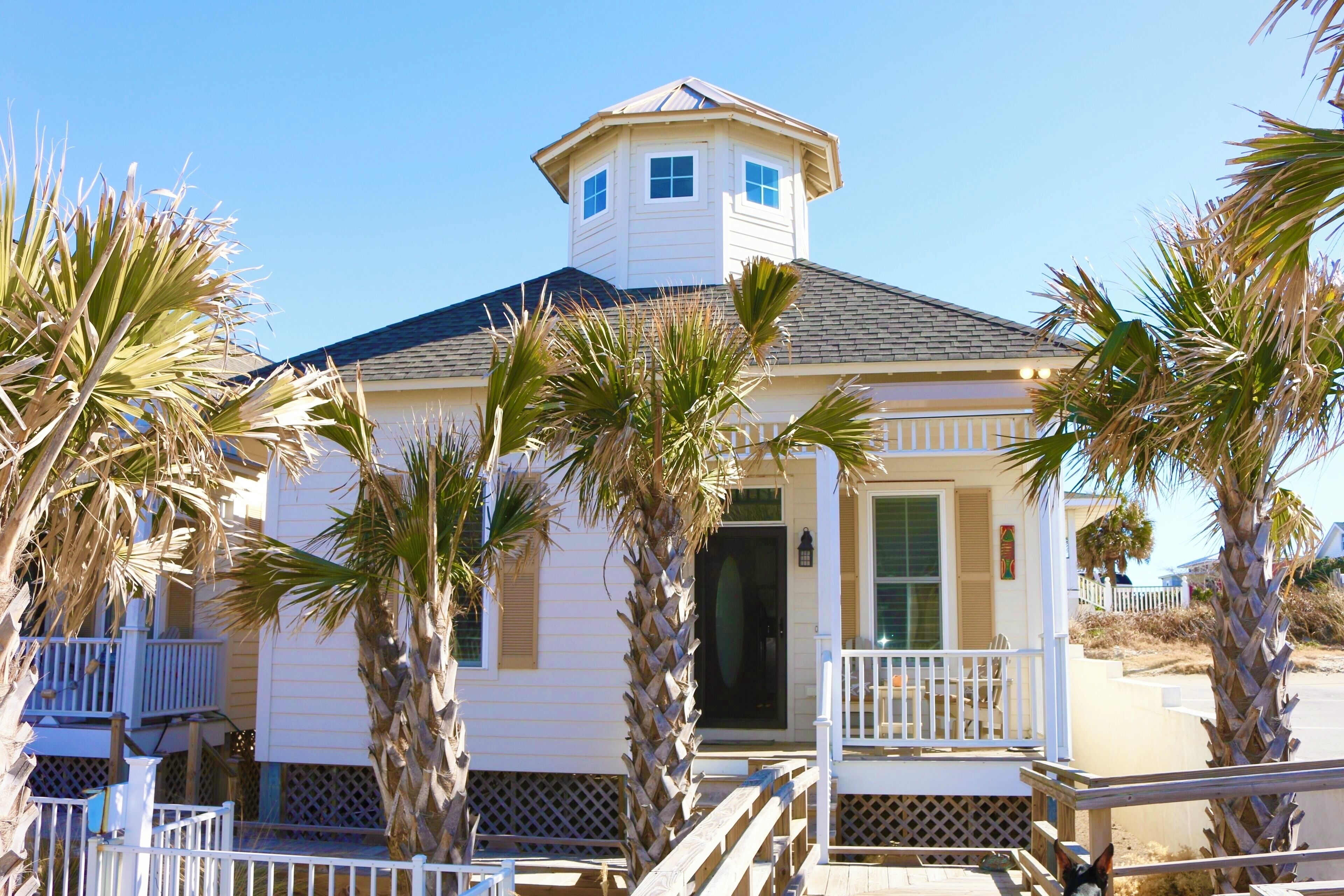 A two-story house with a small tower-like structure on the roof, surrounded by palm trees.