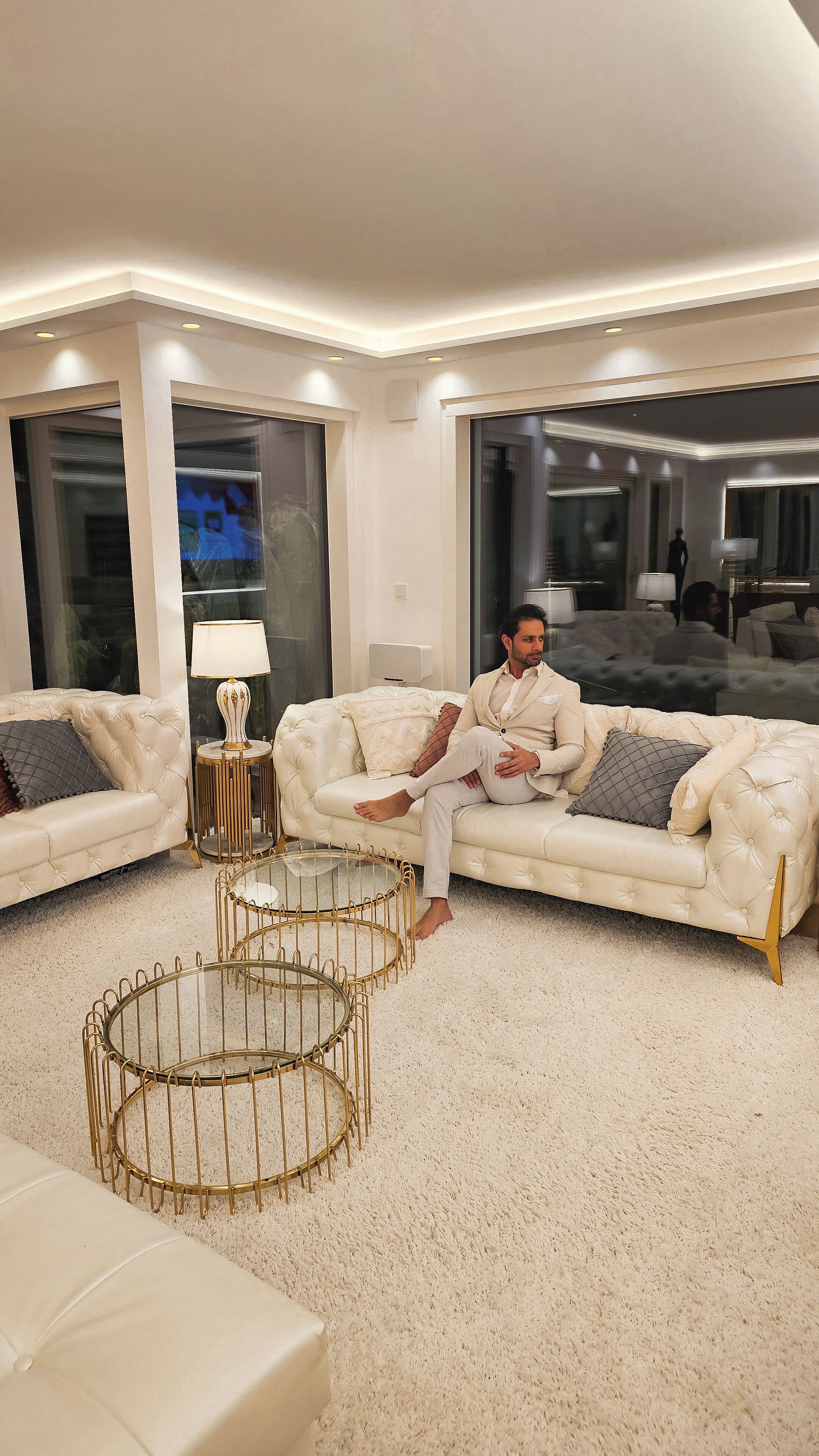 Una sala moderna con un sofá blanco grande, una mesa de centro con marco dorado y un ventanal con vistas a otra habitación.