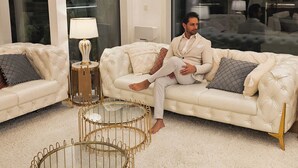 Ein modernes Wohnzimmer mit einer großen weißen Couch, einem Couchtisch mit goldener Rahmung und einem großen Fenster mit Blick auf einen weiteren Raum.