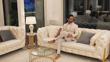 Una sala moderna con un sofá blanco grande, una mesa de centro con marco dorado y un ventanal con vistas a otra habitación.