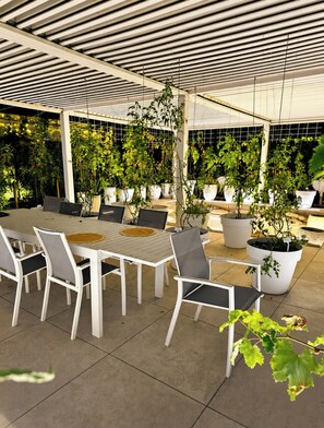 Un patio exterior con una mesa y sillas, rodeado de plantas en macetas y un techo con vigas horizontales.
