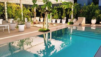 Una piscina con una pérgola cubierta de plantas, rodeada de macetas y un cielo azul despejado.