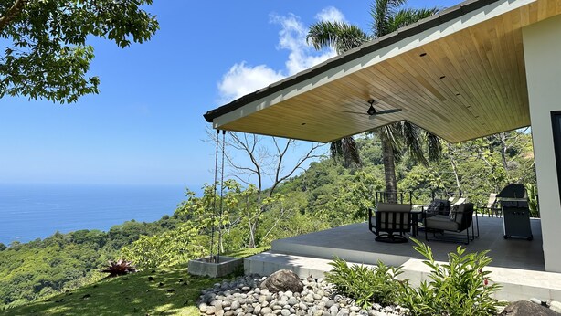 A modern outdoor patio with a ceiling fan, seating, and a grill, overlooking a lush landscape and the ocean. - Fantastic Ocean View Villa (Dominical)