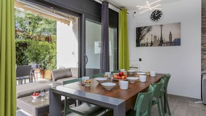 Ein Essbereich mit Tisch für vier Personen, umgeben von grünen Stühlen, einem Glastisch und einer Couch. Ein großes Fenster mit Vorhängen lässt natürliches Licht herein und bietet Blick ins Grüne.