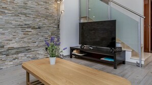 Ein modernes Wohnzimmer mit Steinwand, einer Holzbank, einem Fernseher auf einem schwarzen Standfuß und einer Treppe mit Glastrennläufern.