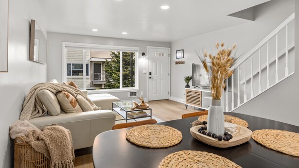 A modern living room with a sofa, coffee table, and dining table. There is a staircase with a railing, a door, and a window with blinds. - Experiencing Edmonds, In the Heart of it All! (Edmonds)