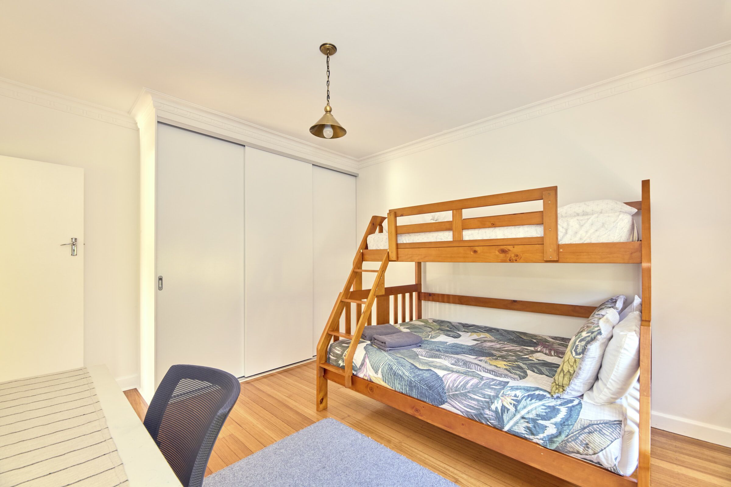 A bunk bed with a wooden frame, a desk, and a chair in a room with white walls and a wooden floor.