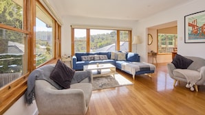 A spacious living room with a large sofa, a coffee table, and two armchairs. There are large windows providing a view of the outdoors. - Basin Road Guesthouse | Walk to Cataract Gorge & CBD (West Launceston)