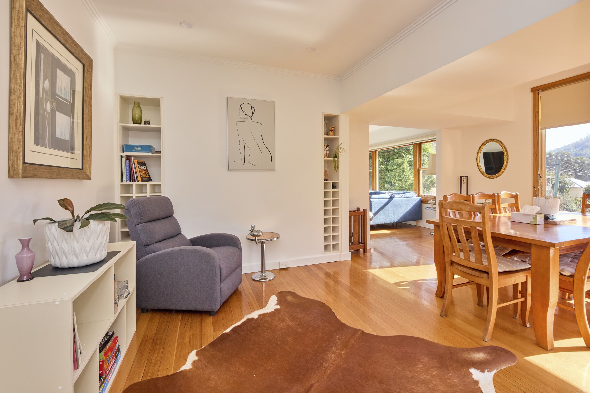 A modern living room with a sofa, a dining table with chairs, a small side table, and a cowhide rug. There are shelves with books and decorative items, a framed picture on the wall, and a window with a view of the outdoors.