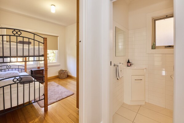 A room with a bunk bed, a small bedside table, a nightstand, a window, a mirror, and a bathroom with a sink and a cabinet. - Basin Road Guesthouse | Walk to Cataract Gorge & CBD (West Launceston)