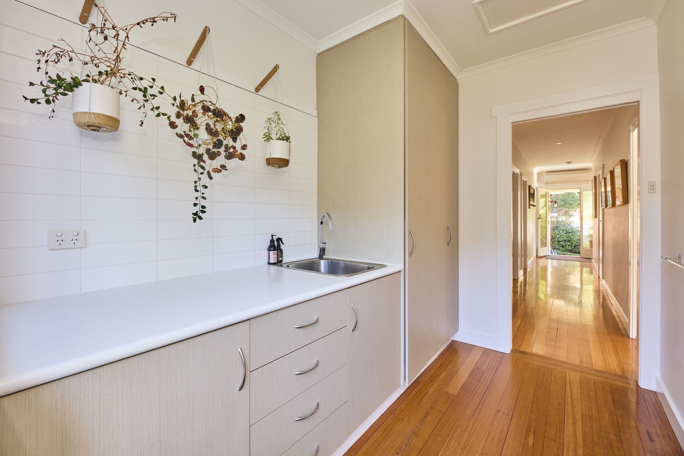 A kitchen with white cabinets, a wooden countertop, and hanging plants. There is a sink with a faucet and a window above it.