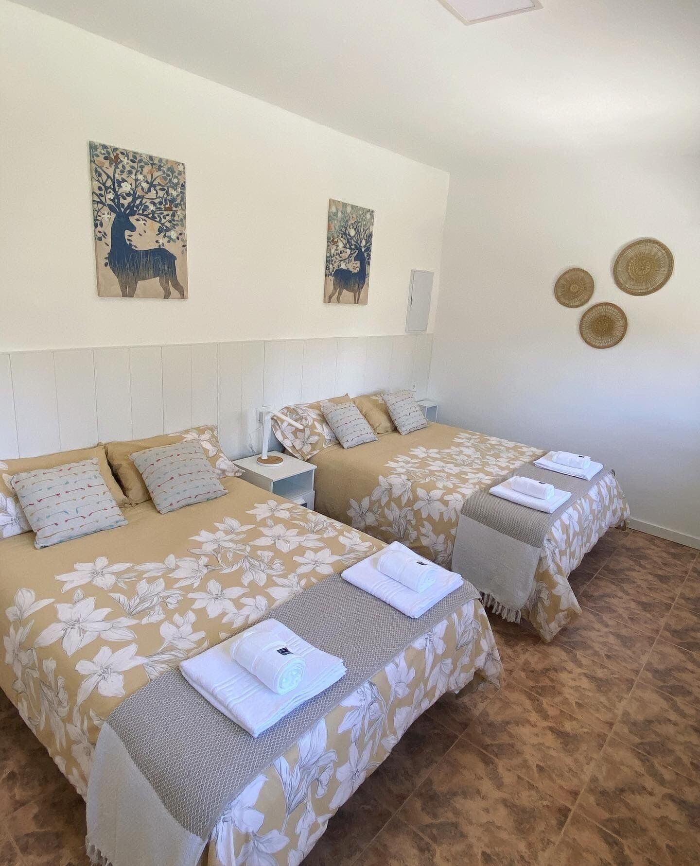 Habitación de hotel con dos camas, cada una con ropa de cama y almohadas florales, una mesita de noche y una obra de arte en la pared que representa ciervos.
