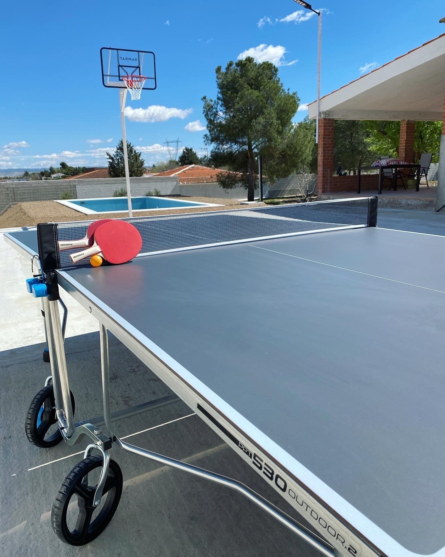 A portable ping pong table with a net, a basketball hoop, and a pool in the background.