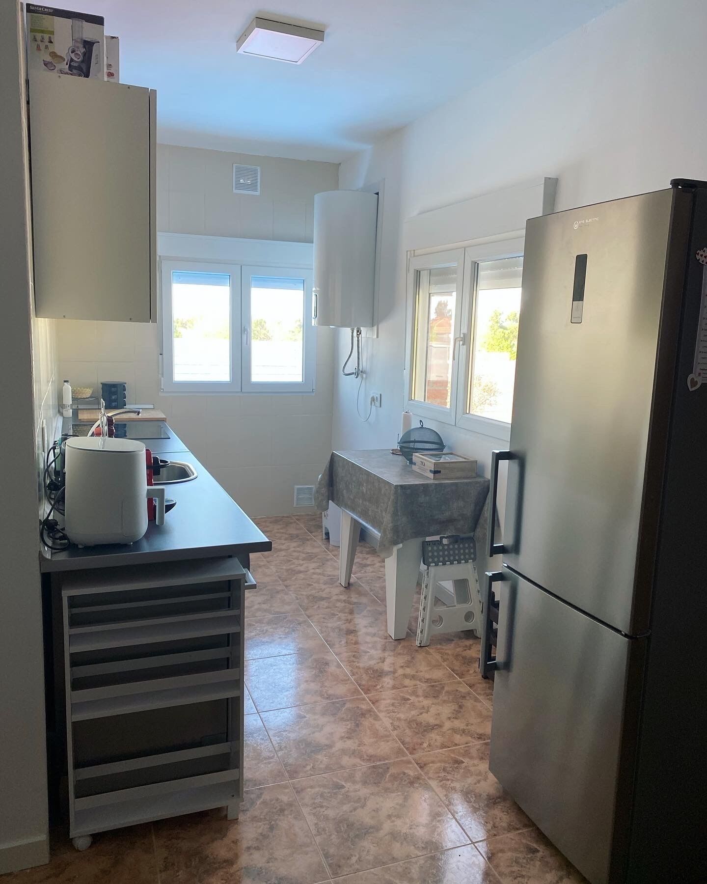 Une cuisine compacte comprenant un réfrigérateur, un plan de travail avec une bouilloire, une petite table et une fenêtre.