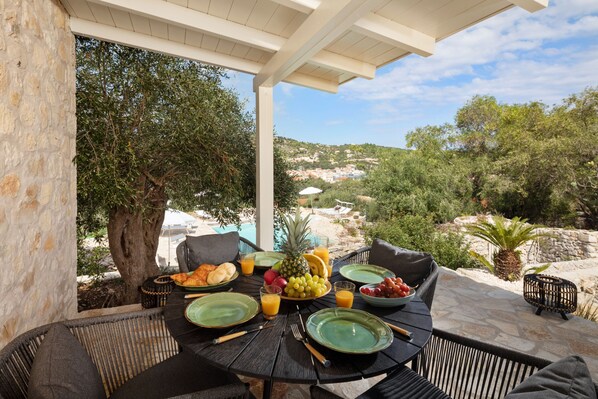 An outdoor dining area with a table set for breakfast, surrounded by stone walls and a view of a hillside.
