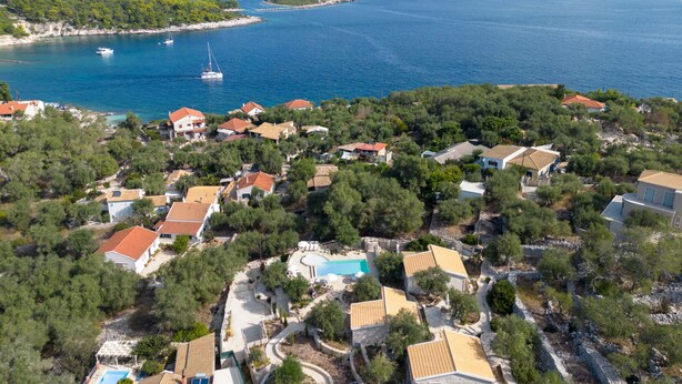 An aerial view of a coastal residential area with houses, a swimming pool, and a clear view of the sea and boats.