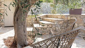 A stone bench and a wicker chair in a stone-walled outdoor area with a tree and a door.