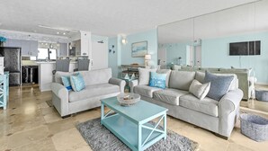 A modern living room with two grey sofas, a teal coffee table, and a kitchen in the background.