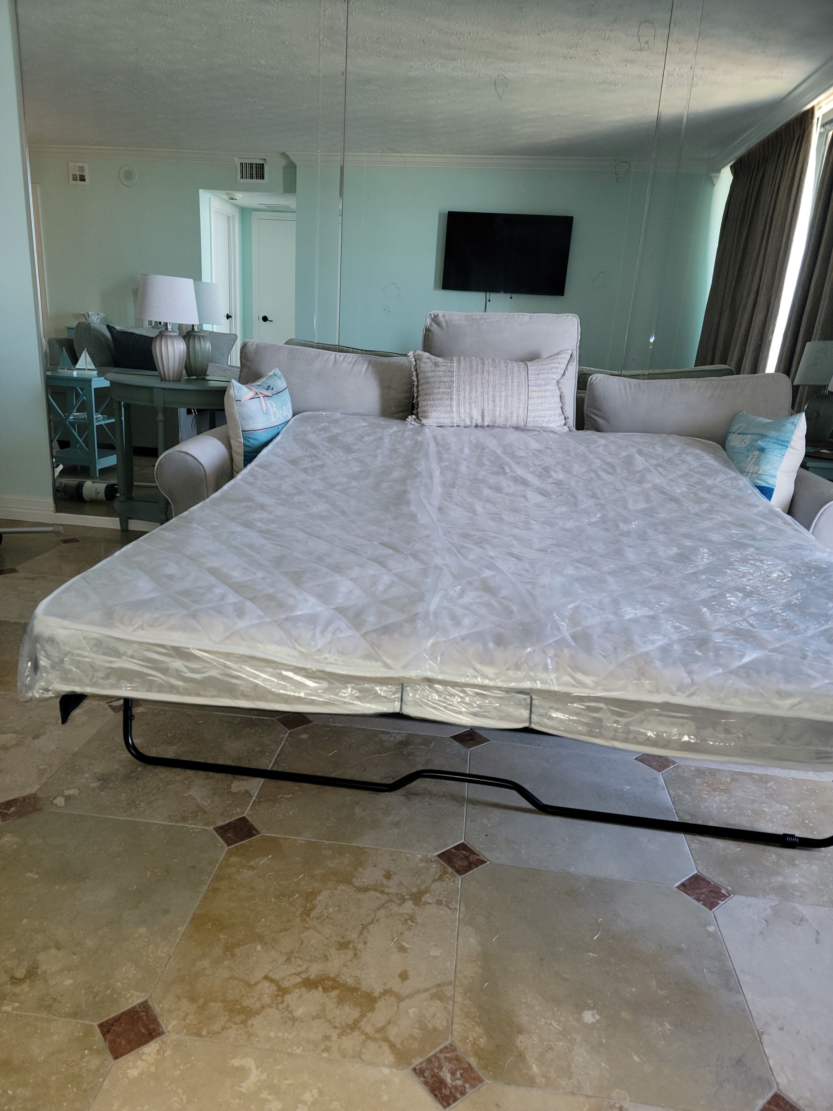 A large bed with a mattress cover, a sofa with cushions, a television on the wall, and a desk with a lamp.