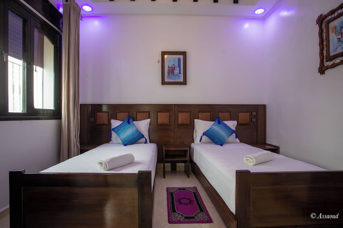 Habitación de hotel con dos camas, cabecero de madera, una mesita pequeña, alfombra morada y un cuadro en la pared.
